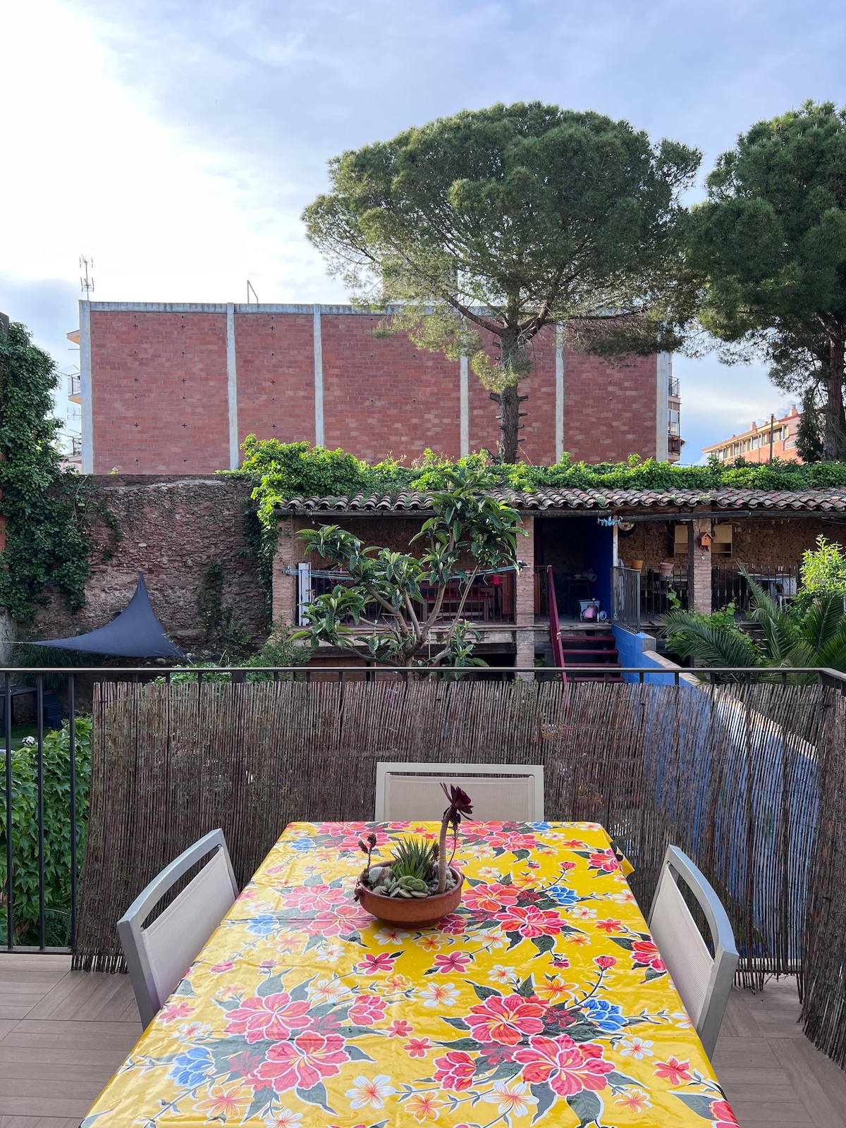 A spacious terrace is displayed, featuring a colorful floral tablecloth atop a wooden table. A small potted plant sits in the center. Lush greenery surrounds the area, creating a natural setting, while a brick building is visible in the background.