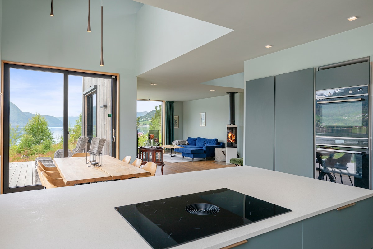 An open-concept living space is featured, showcasing a modern kitchen island with a sleek cooktop. Beyond, a dining table is positioned adjacent to a cozy seating area with a blue sofa. Large glass doors provide a view of the fjord and mountains.