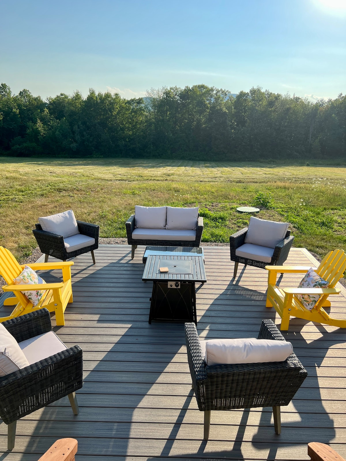 An outdoor patio features comfortable seating arrangements with four black wicker chairs and two yellow Adirondack chairs, placed around a glass-topped table. The area overlooks a peaceful grassy field, framed by trees in the background under a clear blue sky.