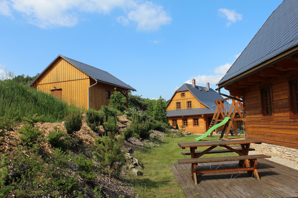 A sunny outdoor area presents wooden chalets surrounded by lush greenery. A picnic table is positioned on a wooden terrace in the foreground, with a playground in the background featuring a slide. The scene captures a peaceful natural setting.