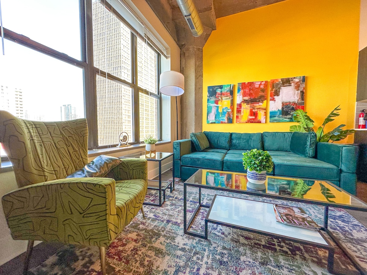 The living area features a vibrant yellow accent wall, complemented by colorful artwork. A spacious green sofa sits alongside a patterned rug, while a green patterned armchair and a glass coffee table add to the modern decor. Large windows provide natural light and city views.