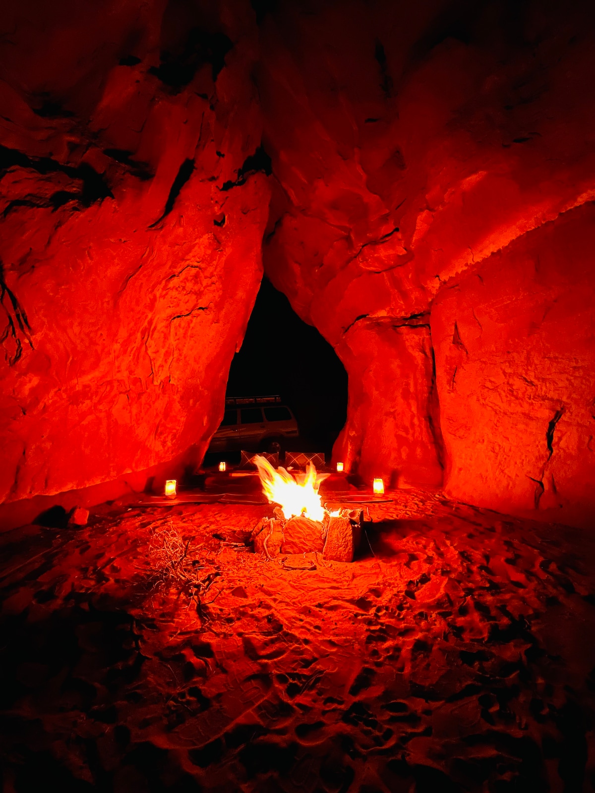 A warm fire burns at the center of a sandy alcove, surrounded by tall, rugged red rock formations. Flickering candles are placed around the area, creating a cozy ambiance. The dark night sky is visible above, enhancing the secluded and serene atmosphere.
