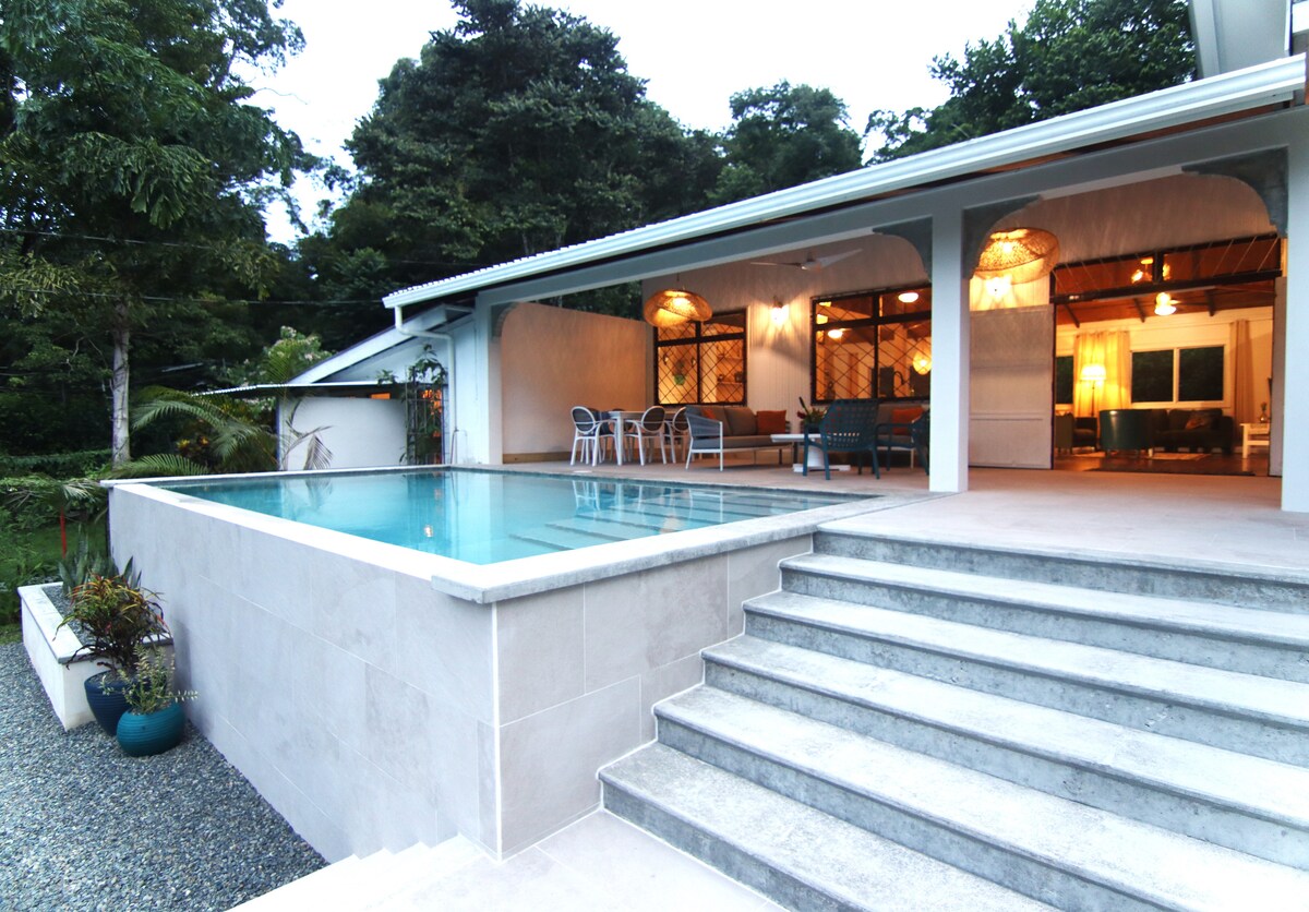 The outdoor area features a swimming pool, adjacent to a spacious terrace with steps leading from the house. The pool area is framed by tropical greenery, with seating options that invite relaxation. Warm lighting can be seen illuminating the interior spaces beyond.