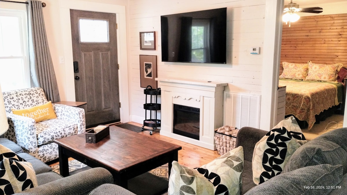 A cozy living room is presented, featuring a comfortable sectional sofa and a patterned armchair. A wooden coffee table is centered, with a decorative box atop it. A wall-mounted TV is positioned above an elegant white fireplace, while a doorway leads to a bedroom in the background.