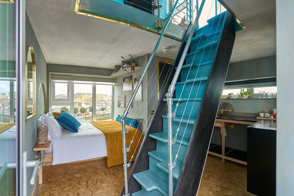 A modern split-level pod interior features a cozy sleeping area with a spacious bed adorned with warm textiles. Distinctive turquoise stairs ascend to the mezzanine level. Large windows allow natural light to illuminate the room, offering views of the surrounding area.