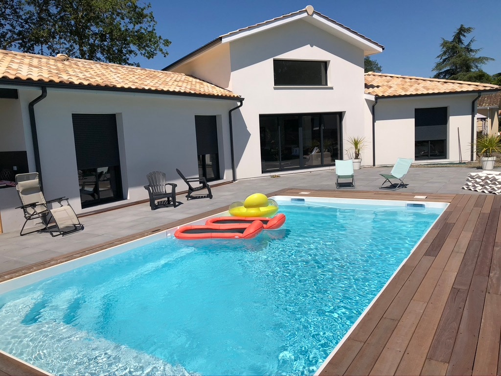 A heated pool is framed by a wooden deck, with lounge chairs positioned nearby. The pool area is complemented by the modern facade of the home, featuring large glass doors that provide a view of the surrounding outdoor space.