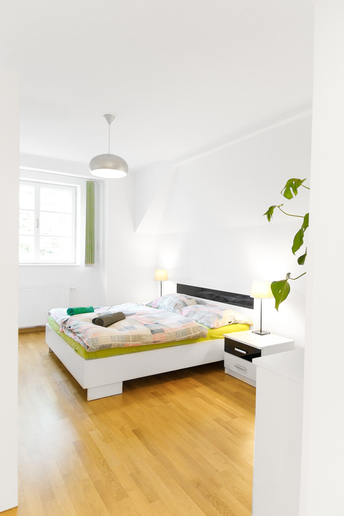 A bright bedroom features a spacious bed with colorful bedding and two bedside lamps providing gentle lighting. Windows allow natural light to fill the room, while a small plant adds a touch of greenery. The wooden floor enhances the room's inviting atmosphere.