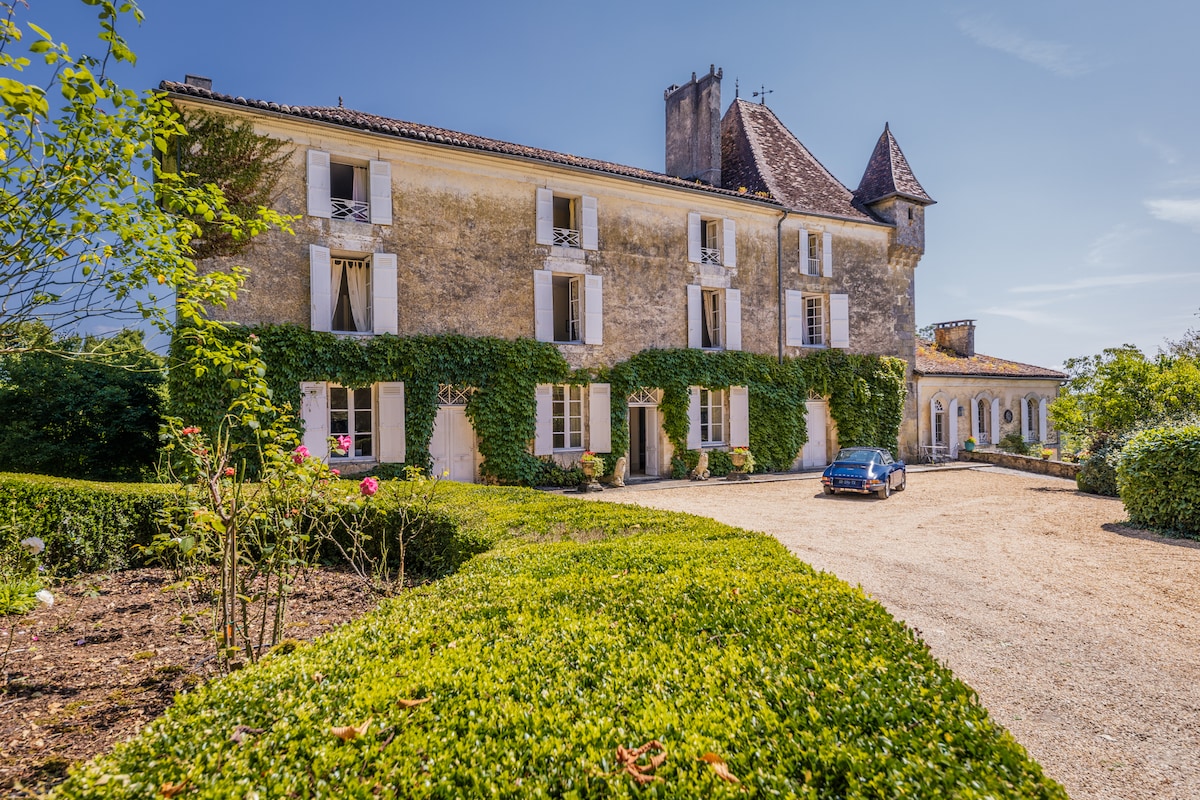 The exterior of a historic chateau is showcased, featuring a stone facade with ivy climbing up the walls. A gravel driveway leads to the entrance, flanked by neatly trimmed hedges and vibrant greenery. A classic car is parked, emphasizing the charm of the property.