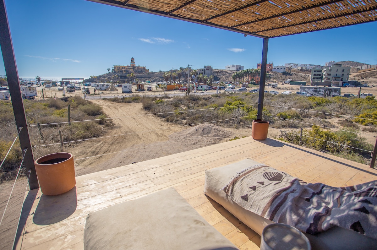 An outdoor seating area is depicted, featuring large cushions on a wooden deck under a thatched roof. A view of sandy paths and a distant hillside town with various buildings is visible, framed by sparse desert vegetation and a clear blue sky.