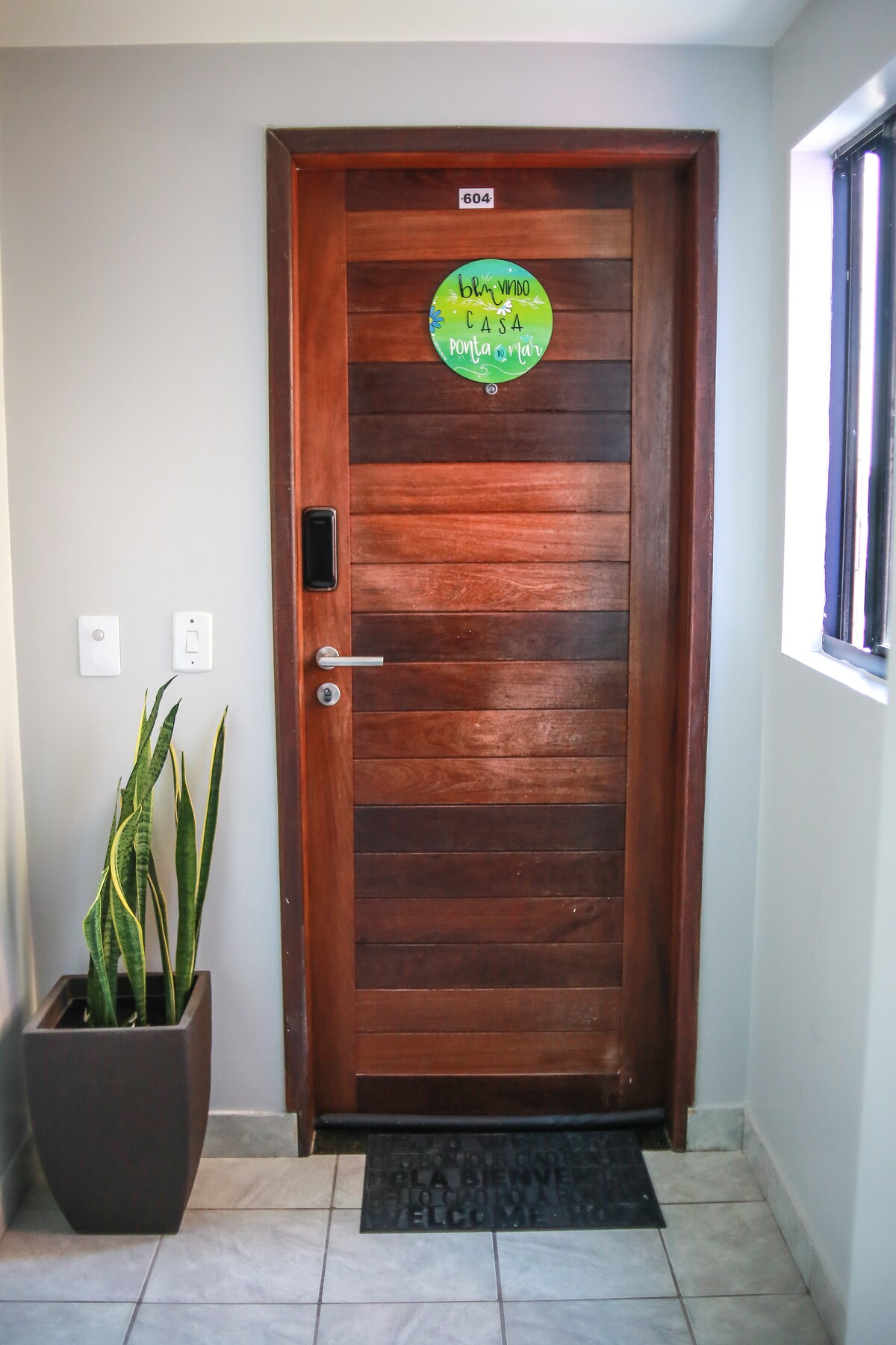 A solid wooden door with a circular green welcome sign is prominently featured. Next to the door, a tall, slender plant in a modern planter adds a touch of greenery. A doormat is positioned at the base of the door, set against a tiled entryway.