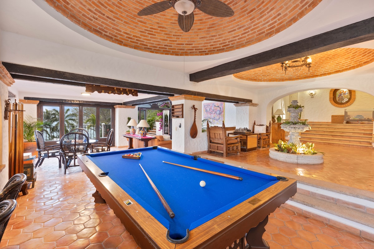 A spacious game area is featured with a blue pool table centrally located, surrounded by natural light from large windows. Brick archways accentuate the ceiling, while decorative elements such as a stone fountain and wooden furniture enhance the inviting atmosphere.