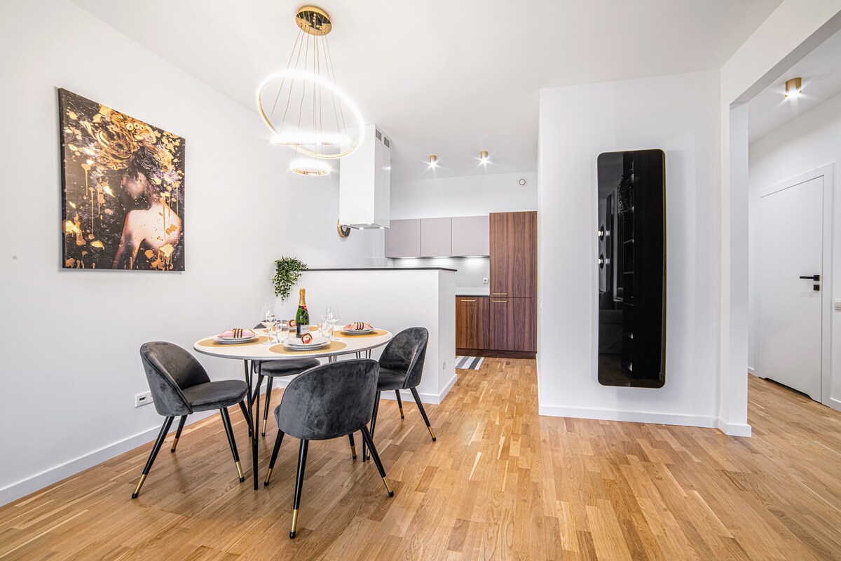 A modern dining area features a round table set for four, complemented by stylish gray chairs. A wall-mounted refrigerator and sleek kitchen cabinetry can be seen in the background. A piece of artwork adds a touch of elegance to the bright, open space.