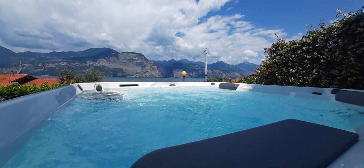 The image captures a hot tub set against a backdrop of mountains and a clear sky. The water appears inviting and is surrounded by lush greenery, enhancing the serene outdoor space with a panoramic view of the landscape.