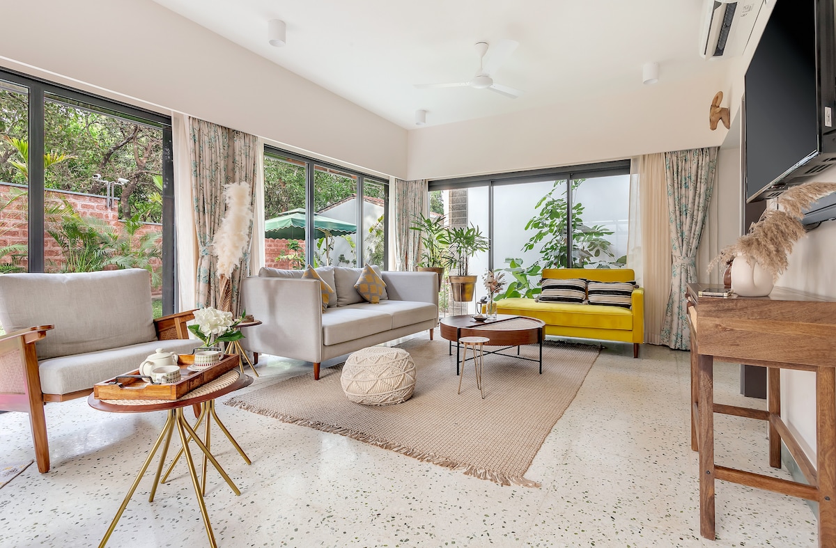 A bright living room is furnished with comfortable sofas and a coffee table, arranged around a soft area rug. Large windows provide views of the greenery outside, with curtains adding a touch of warmth. A flat-screen TV is mounted on the wall, complementing the modern decor.