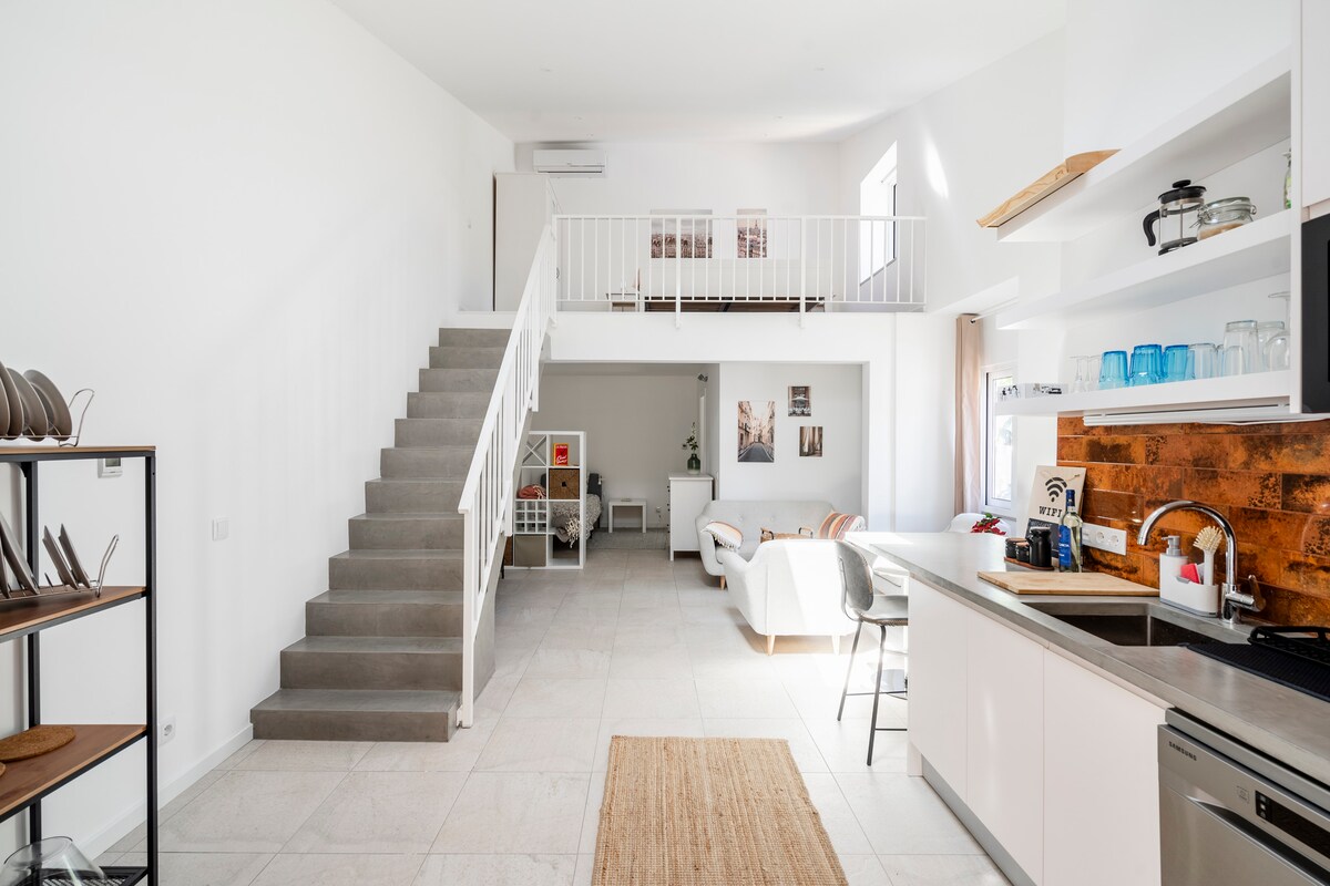 A spacious interior showcases an open-plan layout with a modern kitchen and casual seating area. Bright natural light floods the space through large windows. A staircase leads to an upper level, while a neutral color palette enhances the sense of openness.