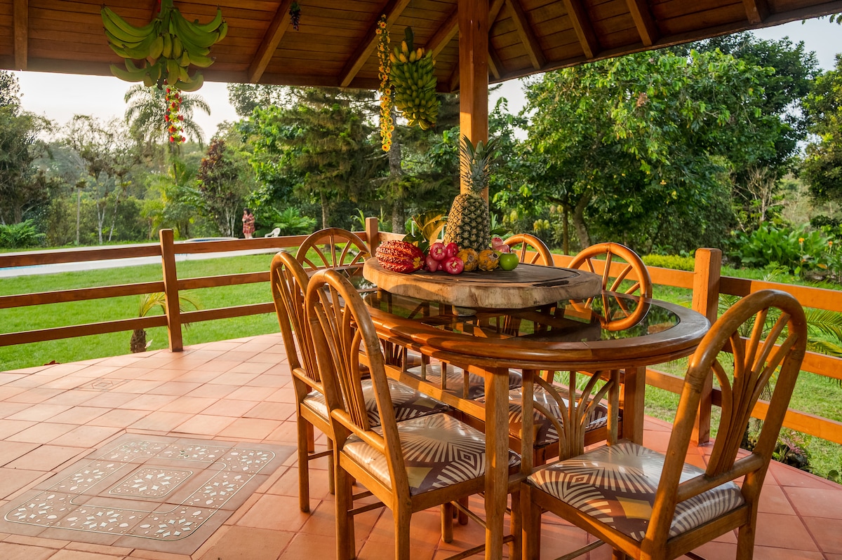 An outdoor dining area features a round wooden table surrounded by six bamboo chairs. Fresh tropical fruits are artistically arranged on the table, with a backdrop of lush greenery and trees, creating a serene environment for outdoor meals.