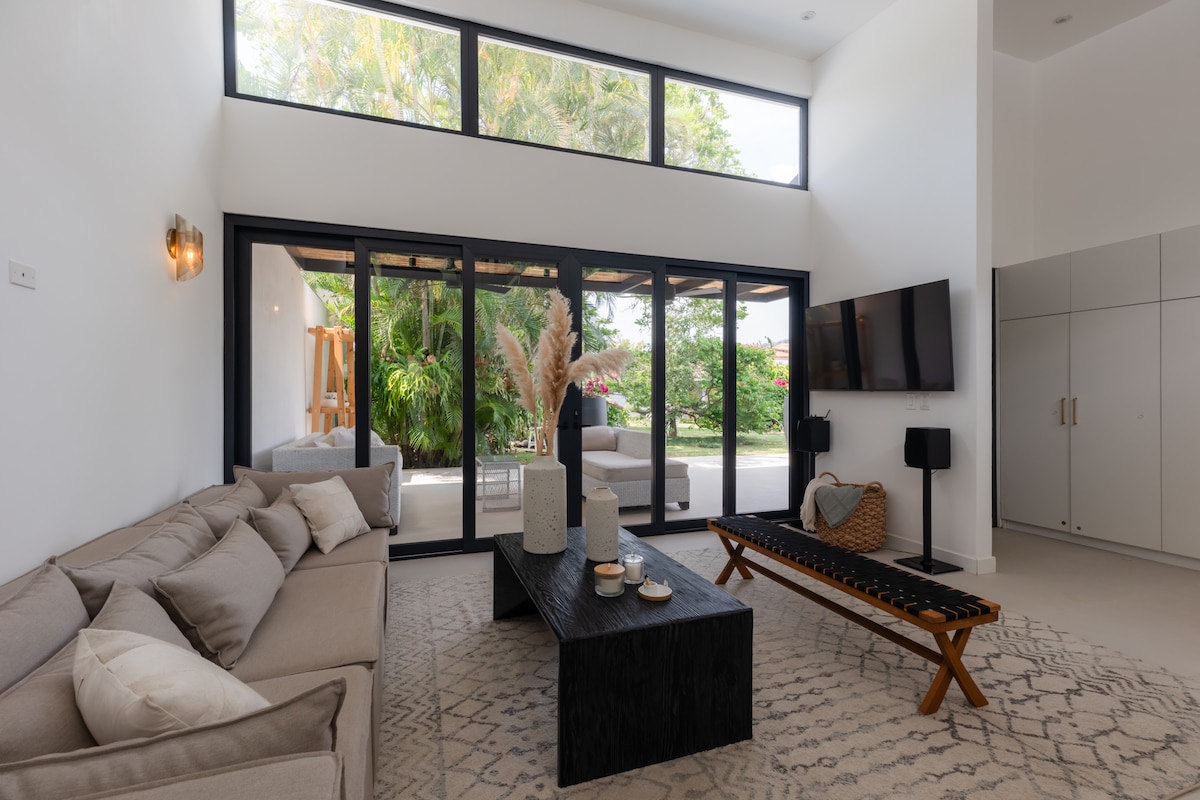 A bright living area is showcased with a large sectional sofa and a textured black coffee table at its center. A flat-screen TV is mounted on the wall across from the sofa. Floor-to-ceiling windows invite natural light, revealing views of the lush garden outside.