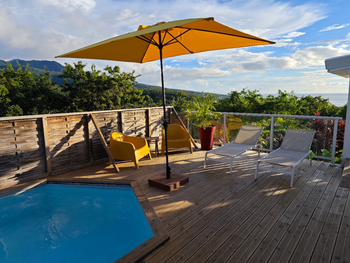A wooden deck surrounds a private pool, featuring two lounge chairs and a bright yellow umbrella. Lush greenery and distant mountains provide a scenic backdrop, enhancing the outdoor space's tranquility.