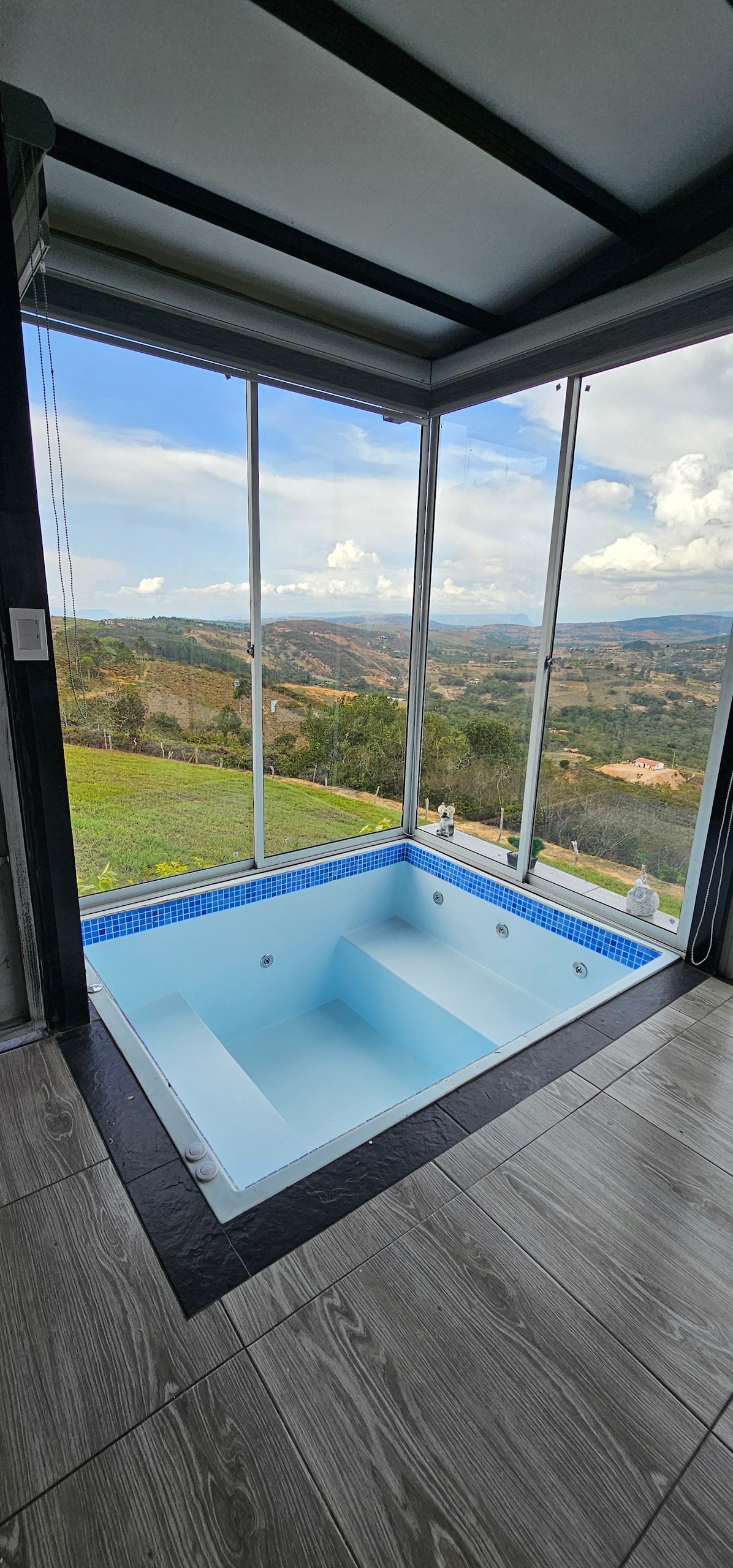 An indoor jacuzzi is positioned beside expansive glass windows, offering sweeping views of the surrounding landscape. The jacuzzi features a clean, light blue interior and is complemented by sleek, dark flooring. Natural light enhances the serene atmosphere.