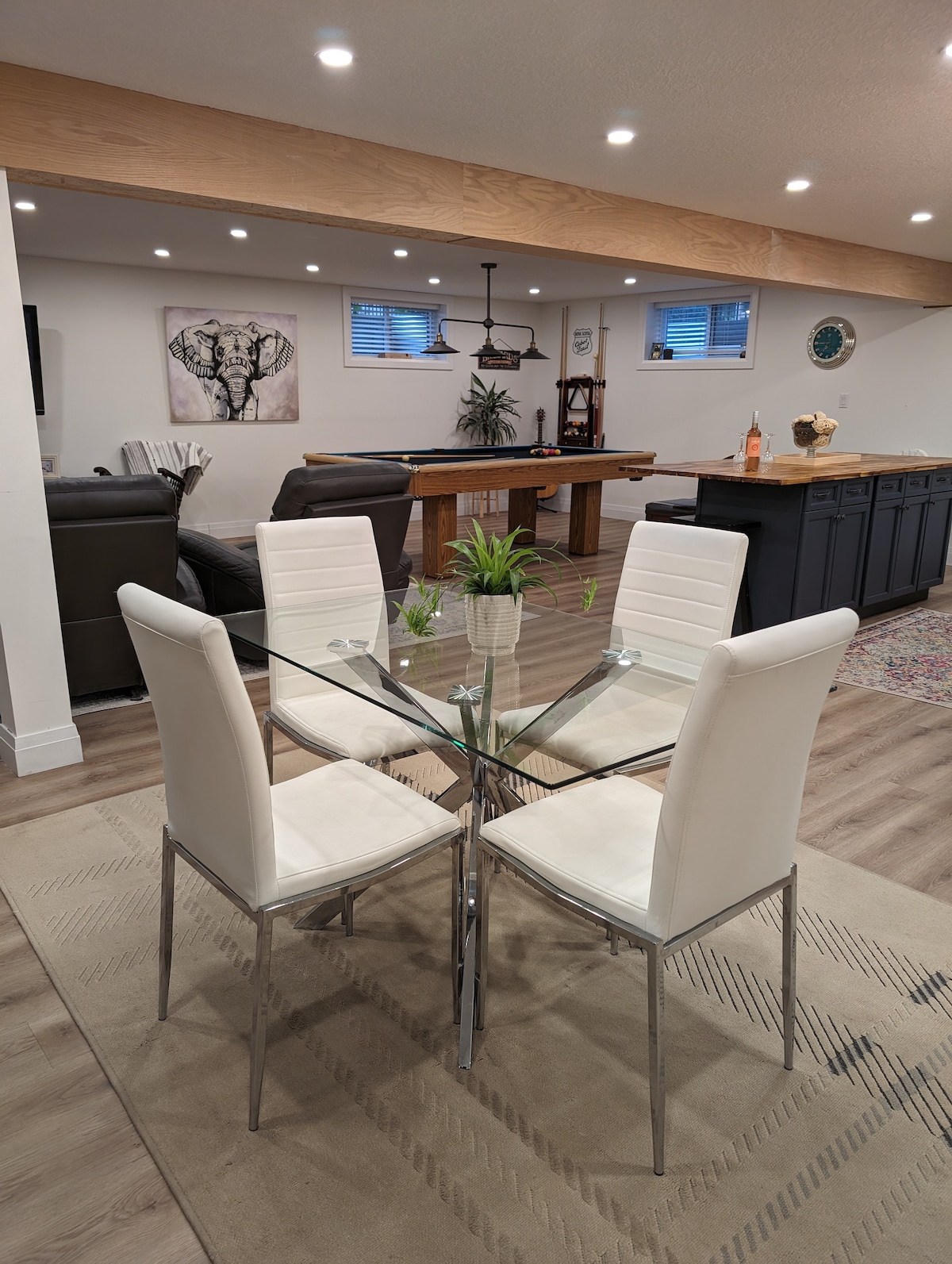 A dining area features a glass table surrounded by four white chairs. The space is illuminated by recessed lighting, and a decorative wall art piece is visible in the background. A plush area rug adds warmth to the wood-like flooring.
