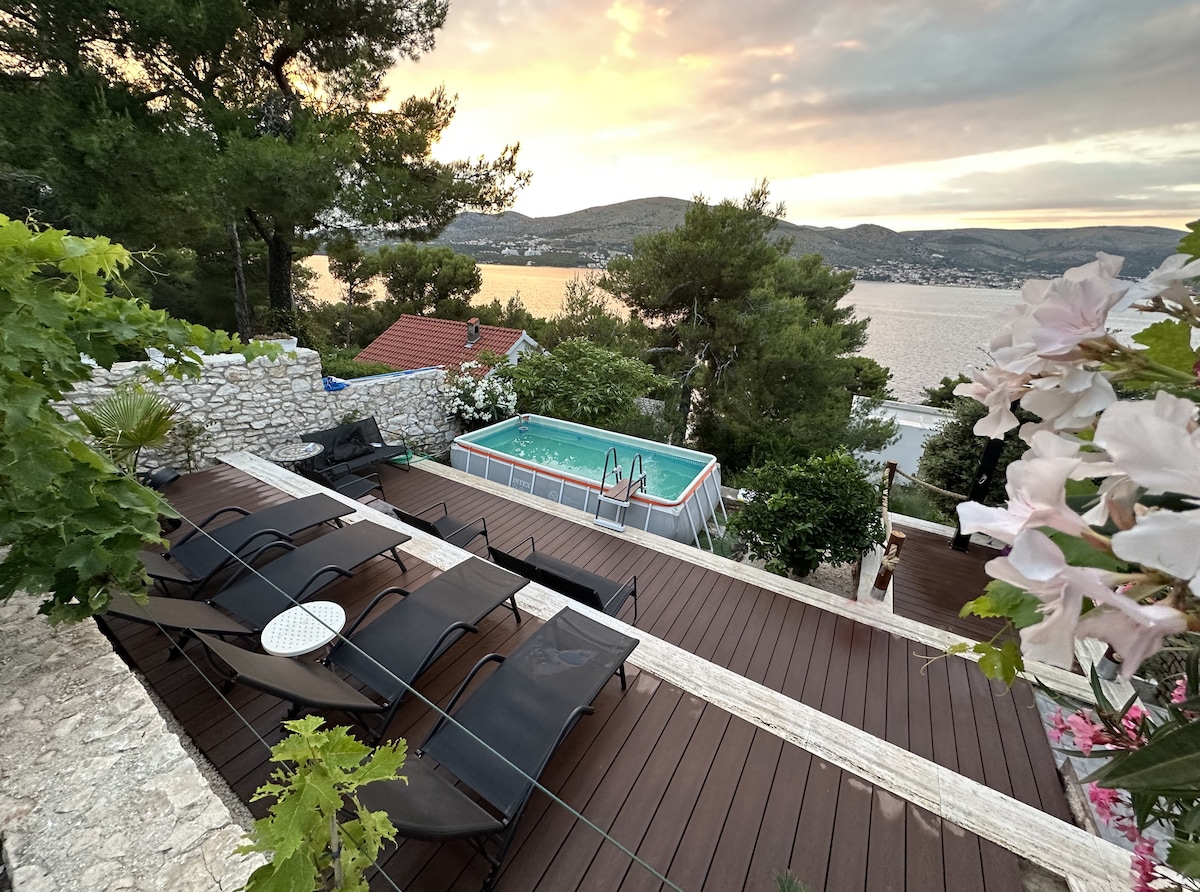 An outdoor terrace showcases lounge chairs arranged around a small, above-ground pool. Lush greenery frames the space, with views of the sea at sunset in the background, creating a serene setting for relaxation and enjoyment.