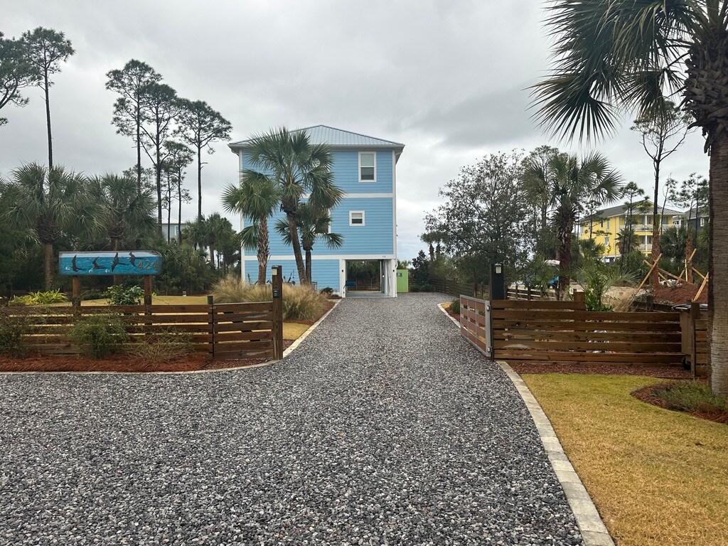 A gravel driveway leads to a two-story blue house, surrounded by palm trees and lush vegetation. The entrance features wooden gates, and the overall landscape reflects a coastal environment with natural greenery.
