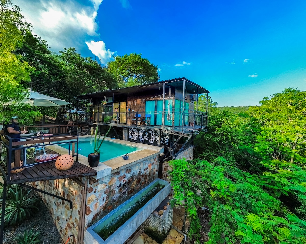 A loft-style house is surrounded by lush greenery, featuring a private swimming pool and a spacious deck with seating. Large windows allow natural light to fill the interior. The structure showcases a modern design with stone accents and a balcony overlooking the scenic landscape.