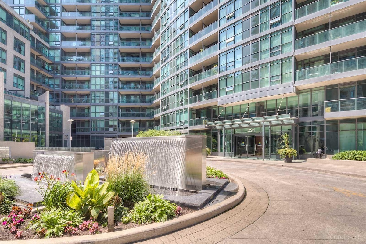 The exterior of the condo building features modern architecture with extensive glass facades. A serene fountain is centrally located in the landscaped area, surrounded by colorful plants and greenery, enhancing the inviting entrance to the building.