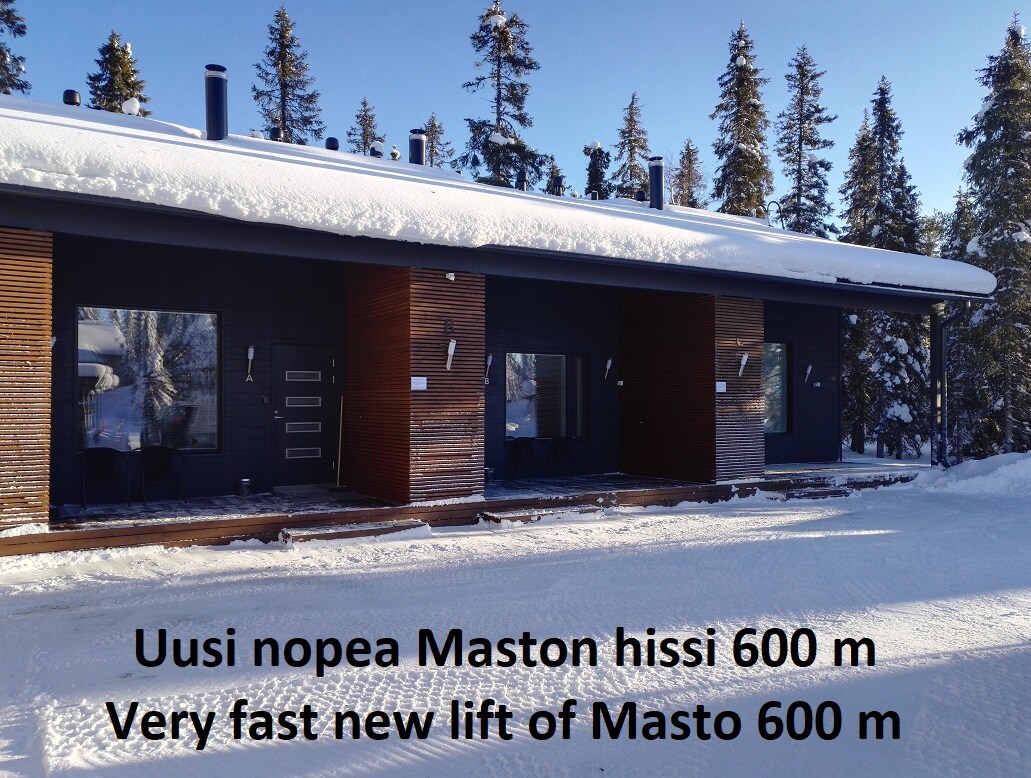 A modern building with a sleek wooden facade is set against a backdrop of snow-covered trees. The entrance area is clear, featuring several doors leading to individual apartments. Snow blankets the ground, indicating the winter season.