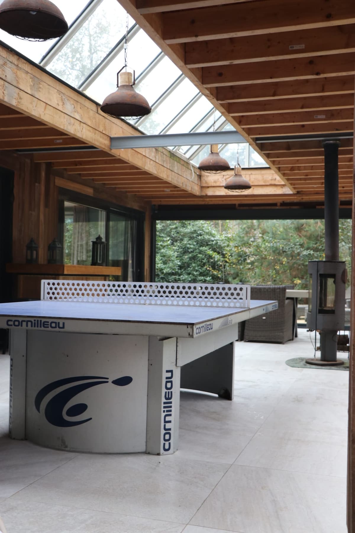 An indoor recreational area features a table tennis table positioned under a glass-roofed section, allowing natural light to fill the space. Wooden beams and pendant lights add to the cozy ambience, while a wood-burning stove provides warmth. Surrounding plants create a serene backdrop.