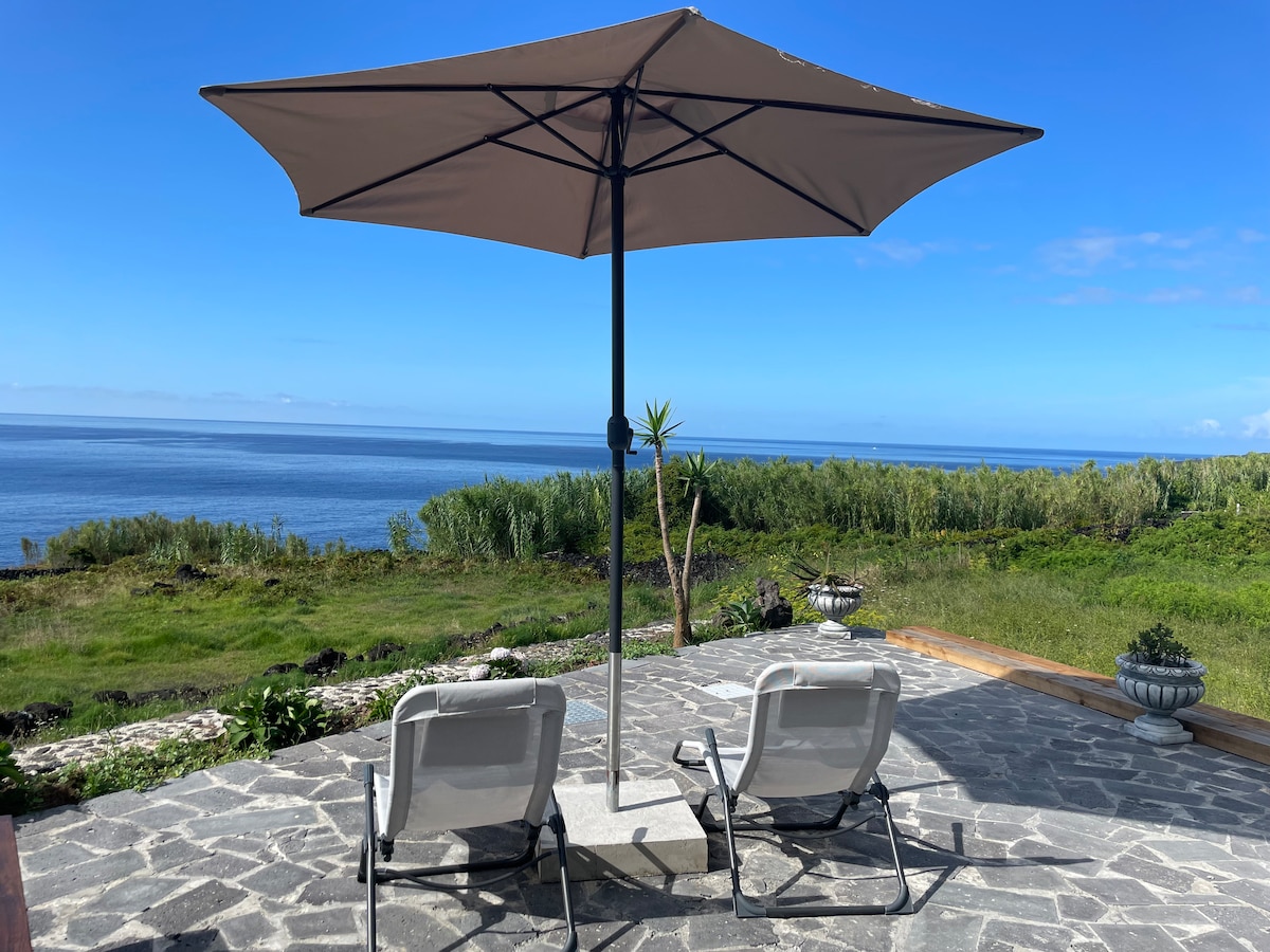 A shaded terrace features two lounge chairs under a large umbrella, overlooking the expansive ocean view. Lush greenery and distant hills contrast with the tranquil blue waters, creating a serene outdoor space ideal for relaxation.
