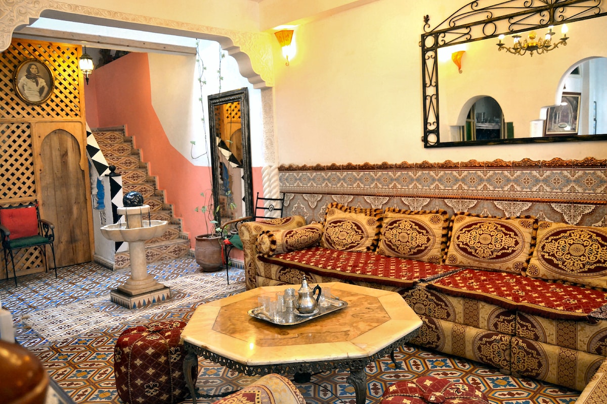 A welcoming lounge area features a large, intricately patterned sofa adorned with cushions, surrounding a low marble table. Colorful tiles cover the floor, and a decorative mirror hangs on the wall. An arched doorway leads to a staircase, enhancing the traditional Moroccan ambiance.