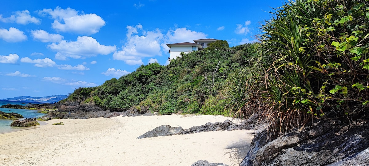 A serene beach scene showcases soft, white sand bordered by rocky formations and lush greenery. The clear blue sky is dotted with fluffy white clouds, while a cozy villa is seen perched on a hillside overlooking the tranquil waters.