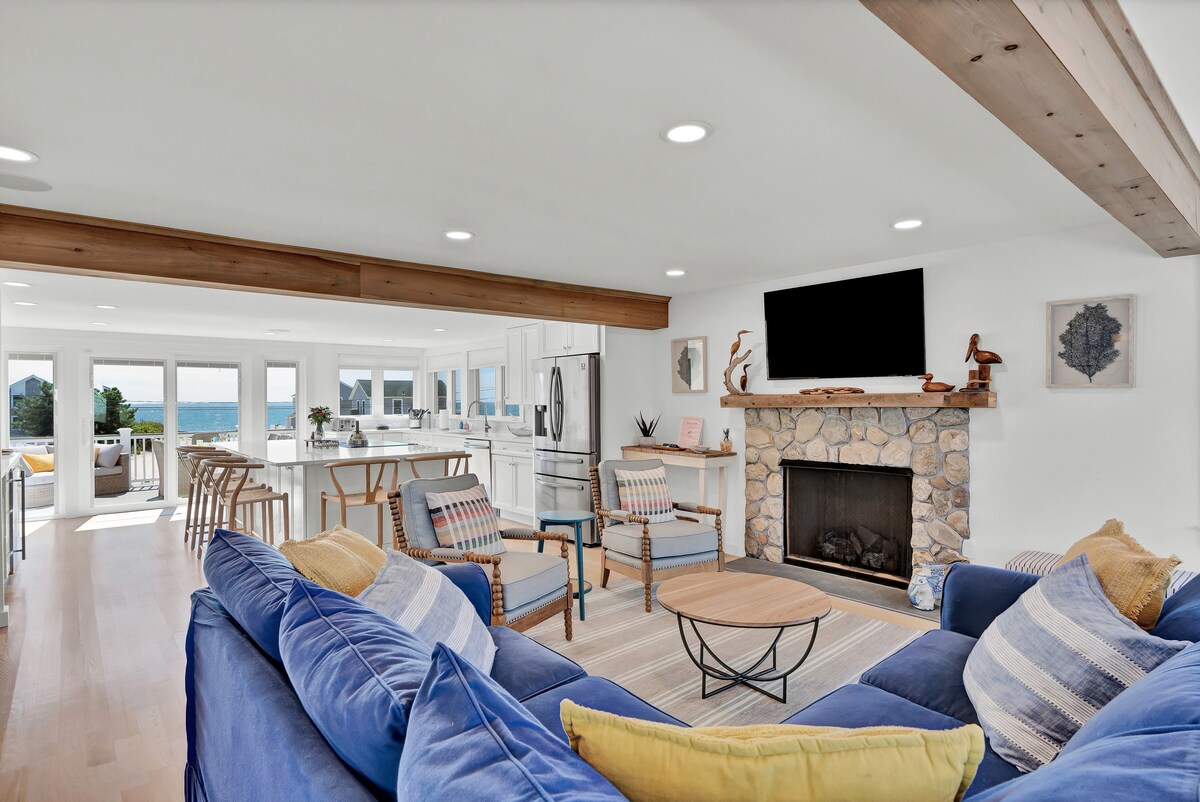 A spacious living area features a comfortable blue sofa set with decorative cushions, situated in front of a stone fireplace. Light streams in through large windows, with a view of the water visible in the background. An adjoining kitchen area is equipped with modern appliances and bar seating.