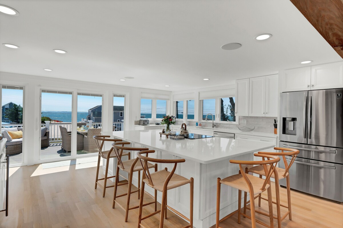 A bright, airy kitchen features an expansive white island with seating. Large windows offer views of the water, enhancing natural light throughout the space. Sleek stainless steel appliances and white cabinetry create a modern and uncluttered environment.
