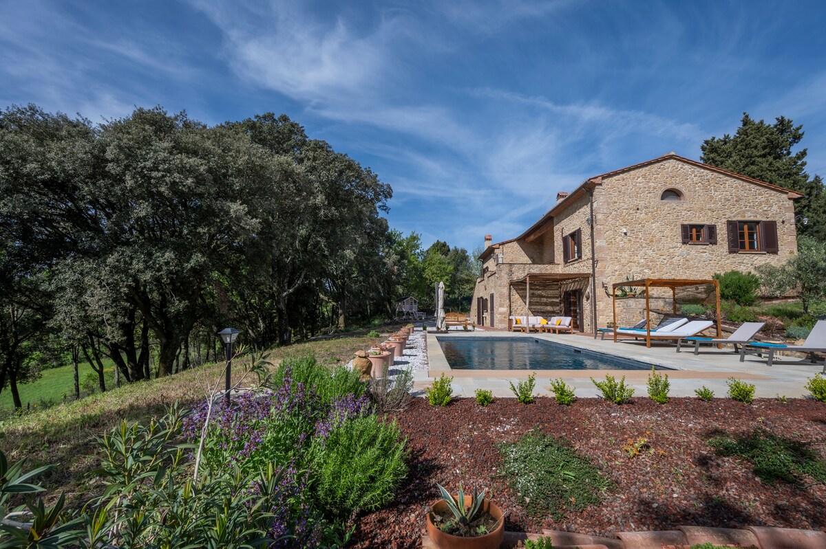 A serene outdoor space surrounds a private pool, with sun loungers arranged for relaxation. Stone walls of the traditional house complement the lush greenery and colorful plants nearby. The clear sky adds to the tranquil ambiance, creating an inviting atmosphere for outdoor enjoyment.