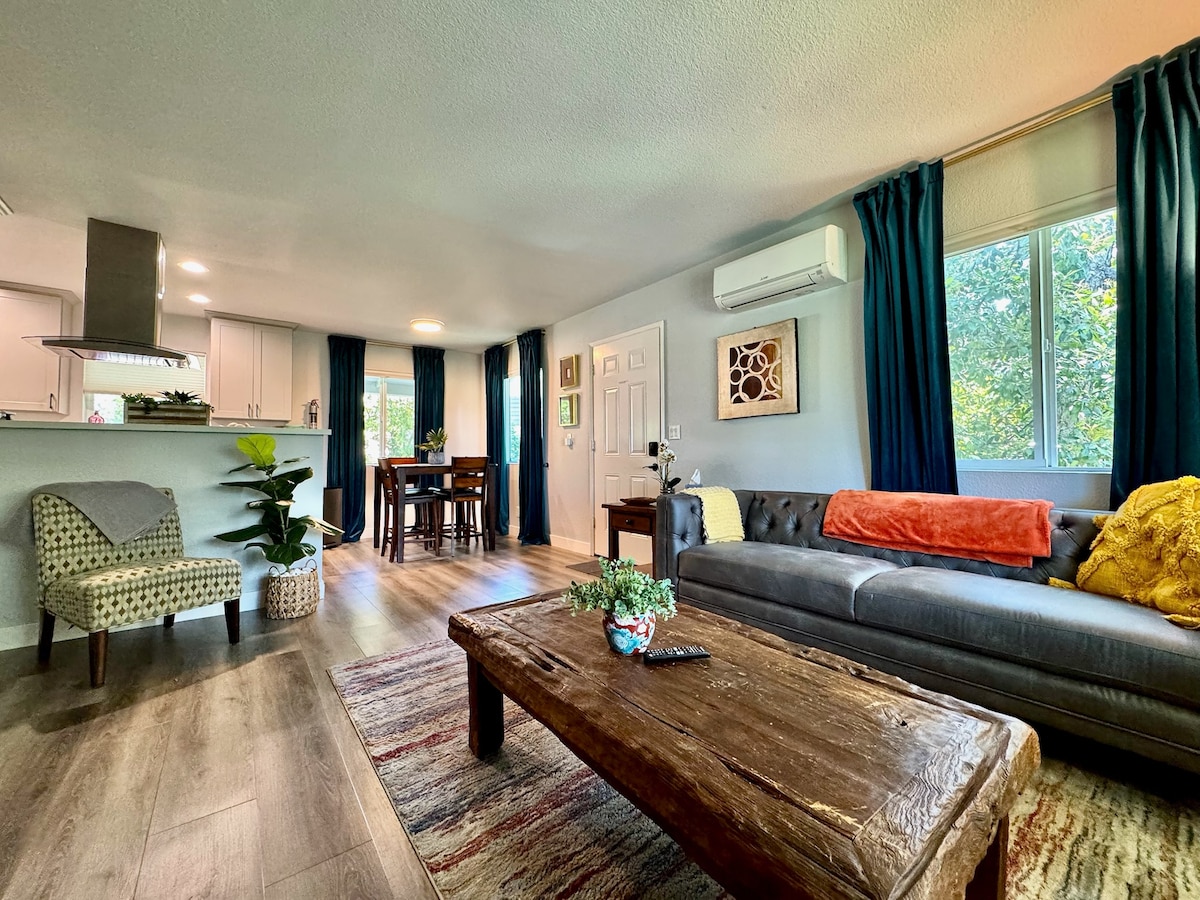 A spacious living area is characterized by natural light streaming through large windows, illuminating the modern decor. A cozy gray sofa is accompanied by colorful cushions, while a rustic wooden coffee table sits in front. A small dining space with a table and chairs is visible in the background.