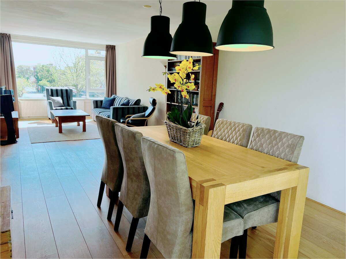 A spacious living and dining area is presented, featuring a large wooden dining table surrounded by six upholstered chairs. Above, three pendant lights illuminate the scene. In the background, comfortable seating is visible near a window, providing ample natural light.
