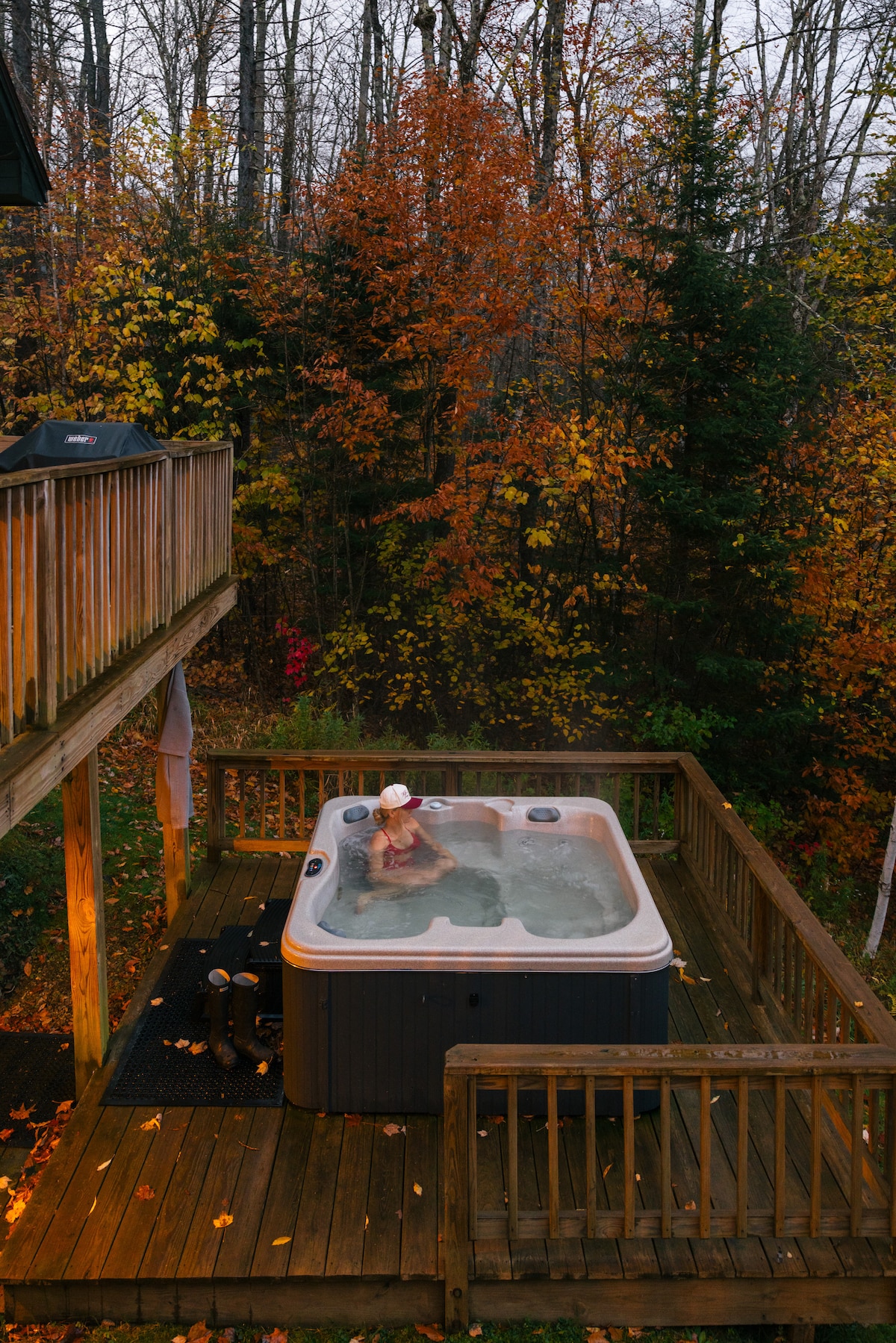 A hot tub is situated on a wooden deck, surrounded by trees displaying autumn foliage. The bubbling water reflects the vibrant colors of the leaves, creating a relaxing environment. A person is seated in the hot tub, enjoying the tranquil outdoor setting.