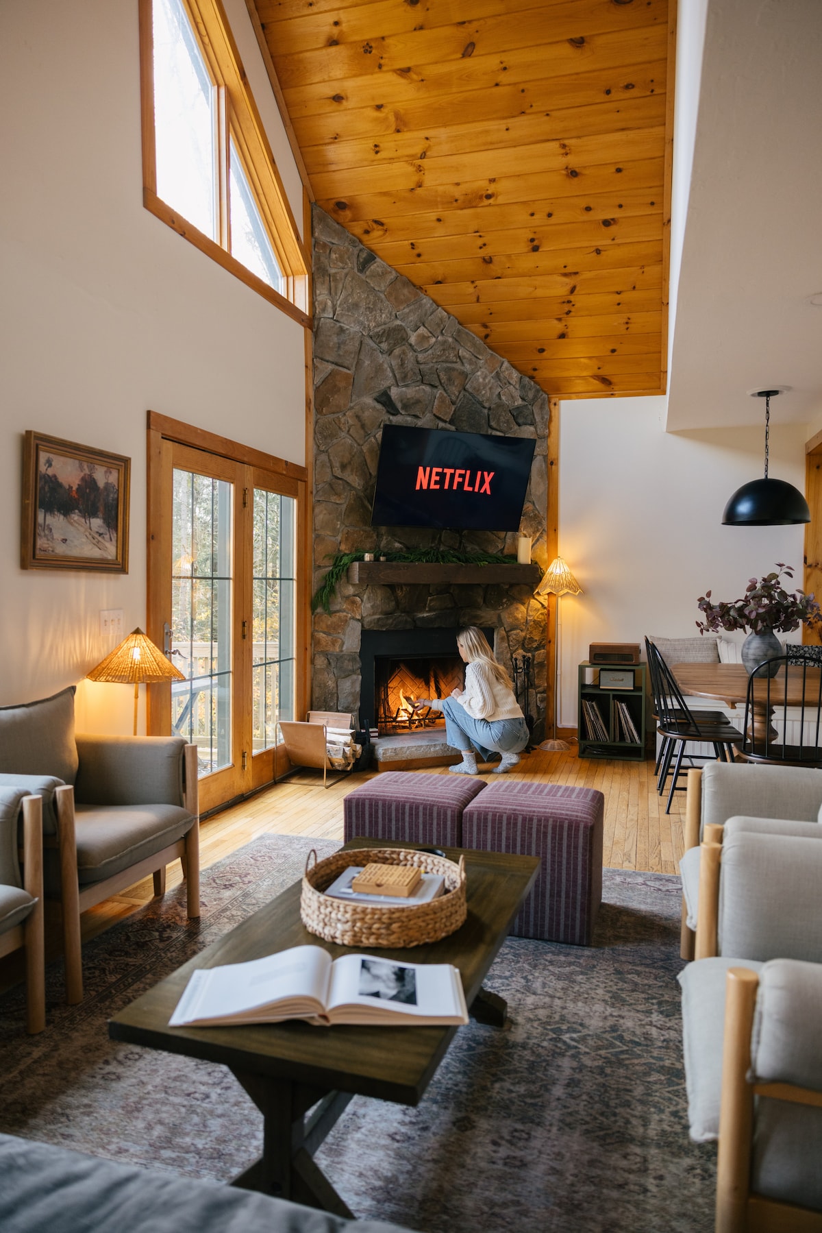 A warm living area features high ceilings and wooden beams. A cozy stone fireplace is flanked by large windows, allowing natural light to fill the space. Comfortable seating is arranged around a coffee table, with a view of a wall-mounted TV displaying Netflix.