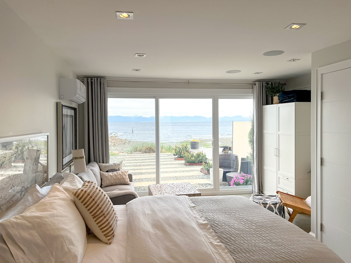 A light-filled studio suite showcases a spacious bed with soft bedding and accent pillows. A seating area is visible beside large glass doors that provide views of the ocean and outdoor space. Plants and flowers add color to the beachside setting.