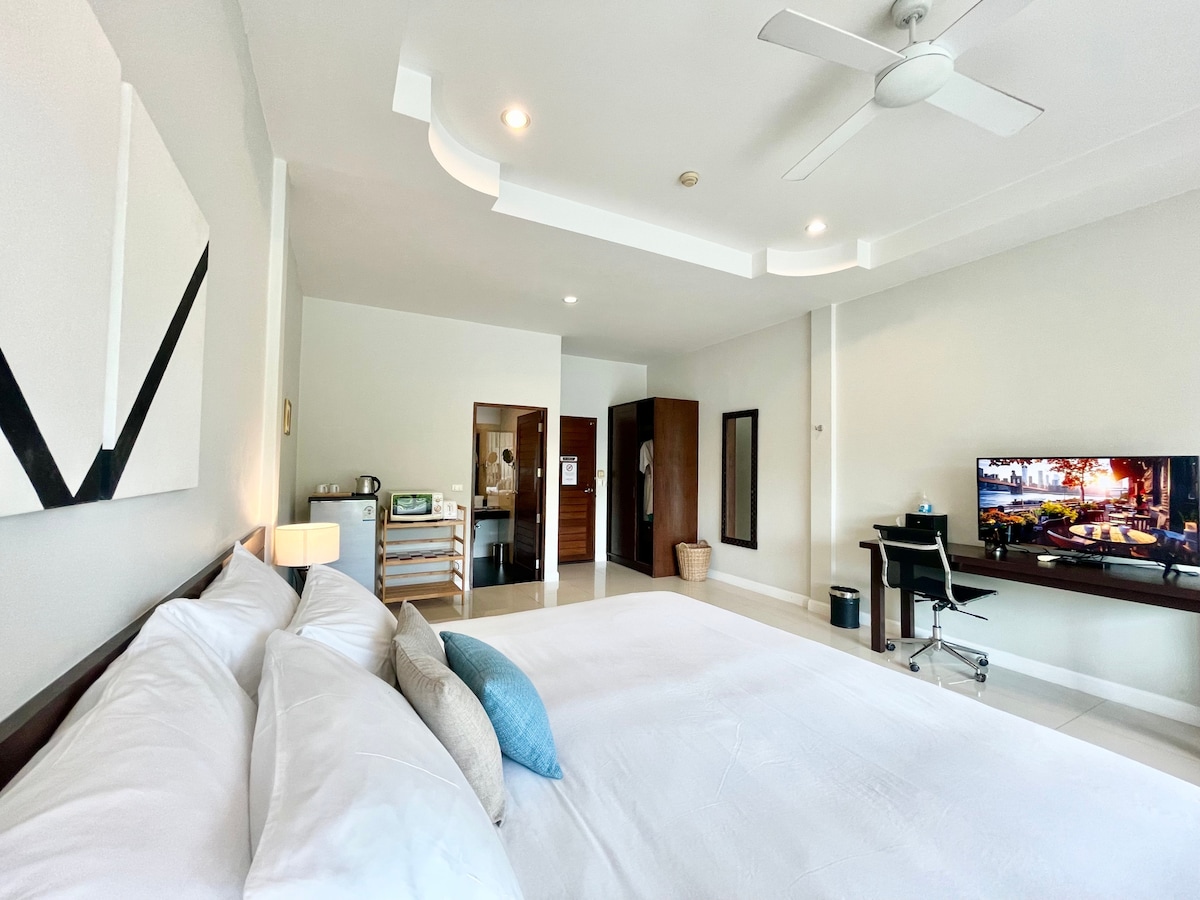 A spacious studio apartment is presented with a large bed adorned with crisp white linens and decorative pillows. A flat-screen TV is mounted on the wall, and a desk with a chair is positioned nearby. Natural light brightens the room, highlighting the modern design.