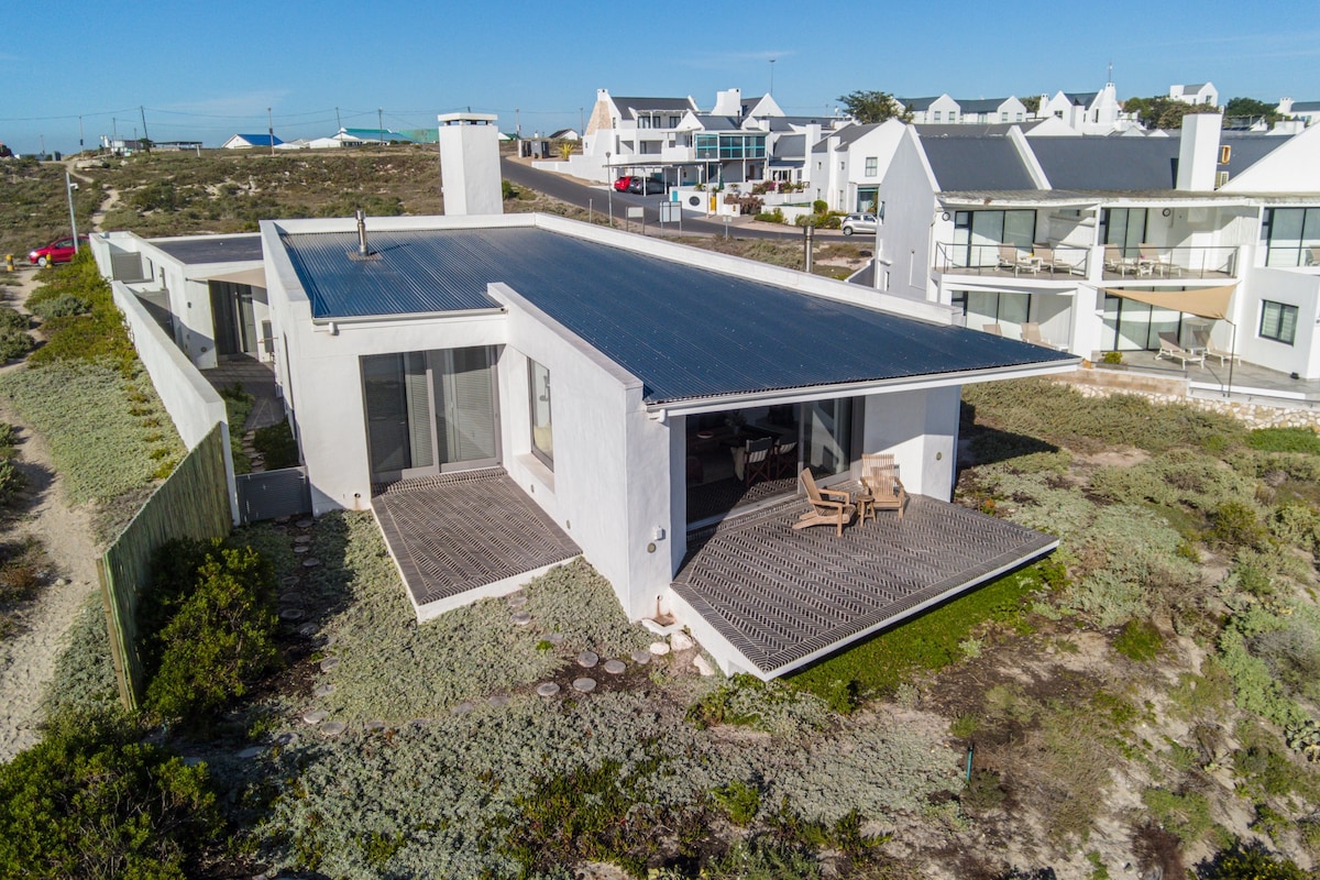 The exterior of the property reveals a modern design with solar panels on the roof. Surrounding vegetation is visible, along with a spacious outdoor area featuring wooden furniture. Large sliding doors are observed, providing access to the interior spaces and promoting a seamless connection with nature.