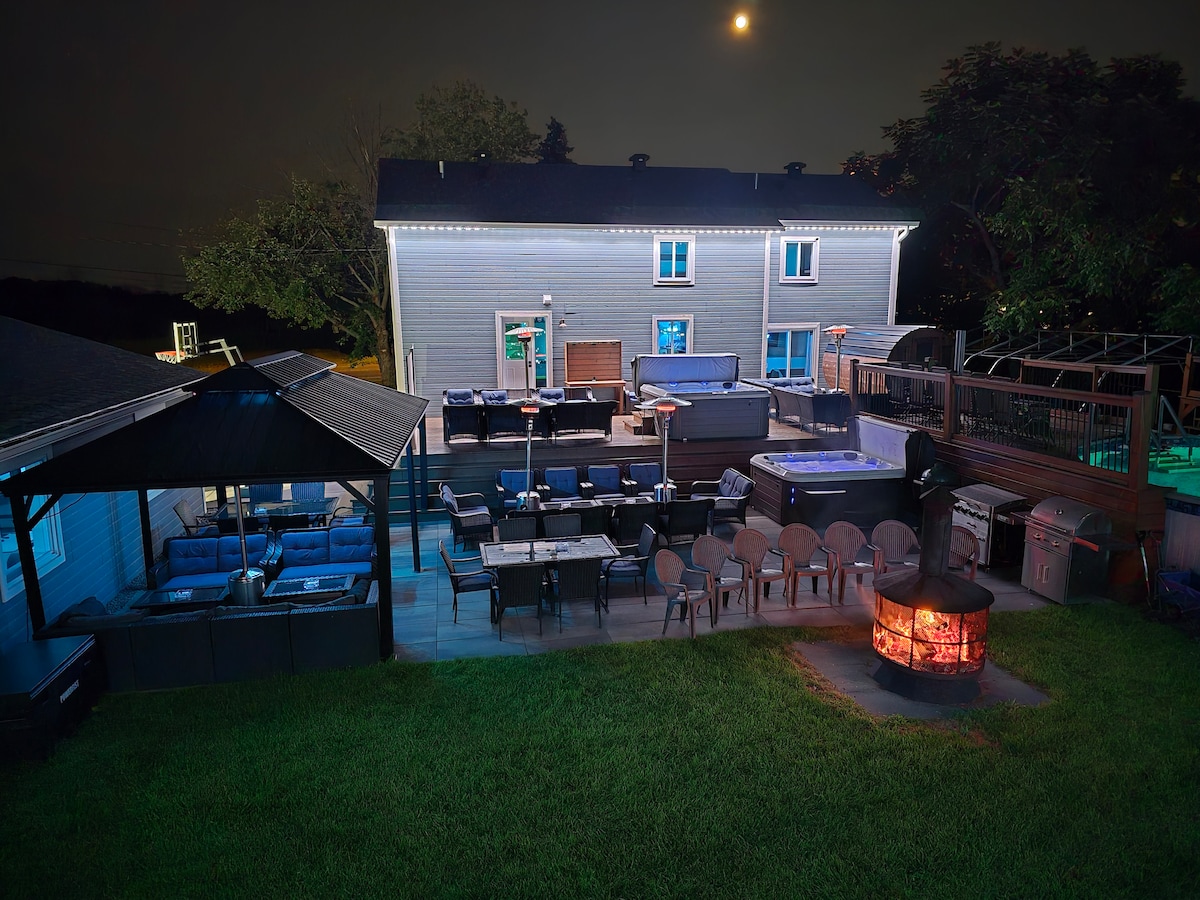 An outdoor space is illuminated under a full moon, featuring a wooden deck with various seating areas. A fire pit is positioned centrally among numerous chairs, while a dining table accommodates several guests. Additional lounge and grilling areas are present, highlighting a spacious and well-equipped backyard.
