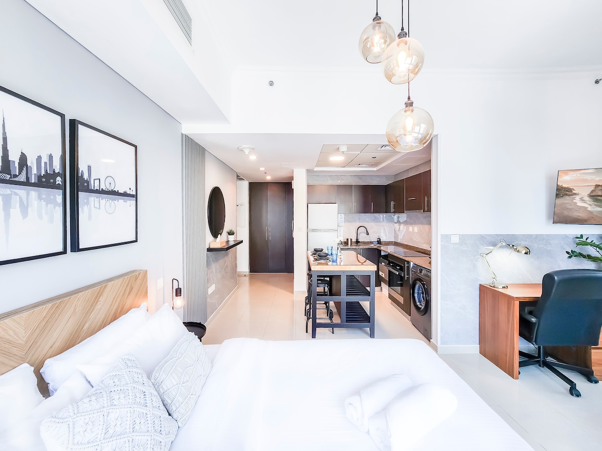 A cozy studio space features a queen-size bed with crisp linens and stylish artwork on the wall. An open-plan living area with modern kitchen appliances and a work desk is visible. Soft lighting is complemented by a large window with city views.