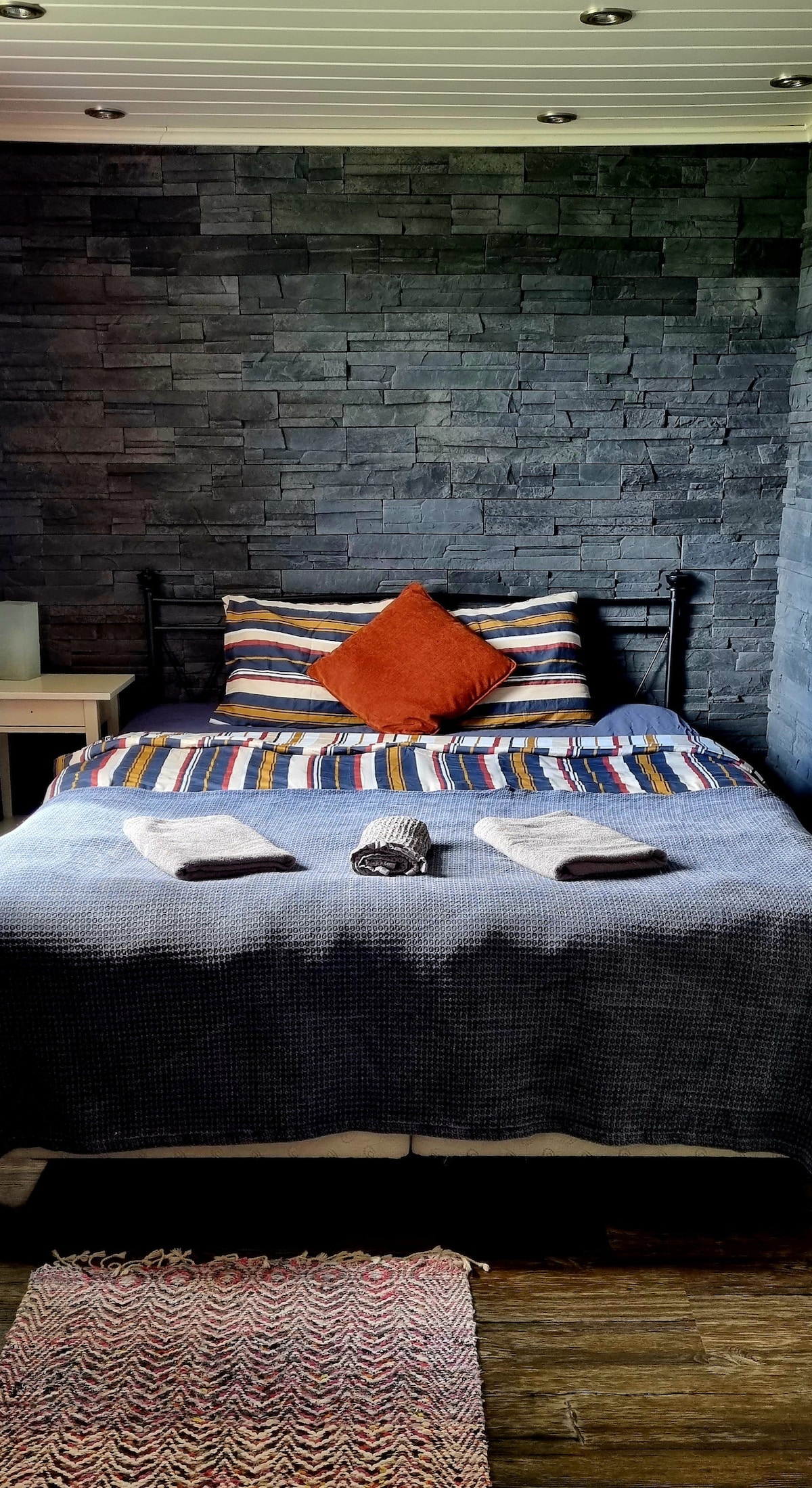 A cozy sleeping area features a bed with a striped blanket and decorative cushions, set against a textured stone wall. Two towels are neatly arranged on the bed, and a woven rug lies on the wooden floor, adding a touch of warmth to the space.