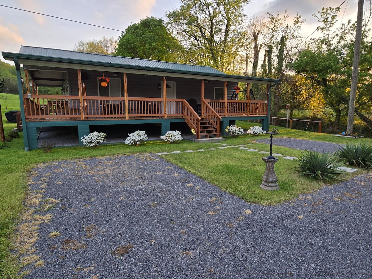 The cabin features a welcoming facade with a green roof and a large wrap-around porch, surrounded by well-maintained landscaping. Rocking chairs are placed on the porch, ideal for enjoying the outdoors. A gravel driveway leads to the entrance, enhancing accessibility to the property.
