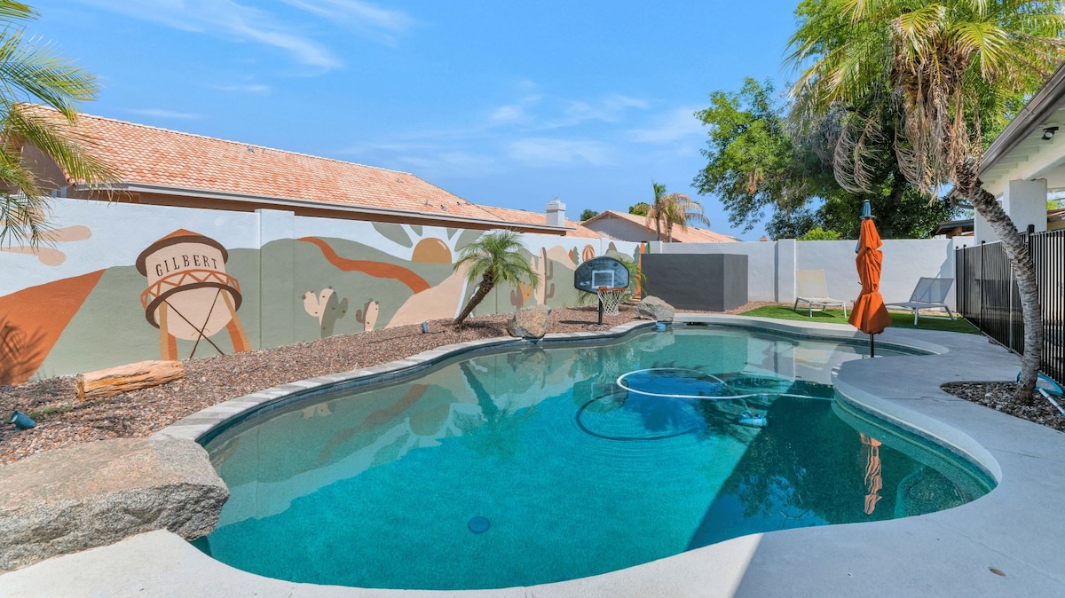 A sparkling pool is set against a vibrant mural depicting the Gilbert landscape, with palm trees lining the edge. The pool area features a basketball hoop, surrounded by stone accents and decorative gravel.