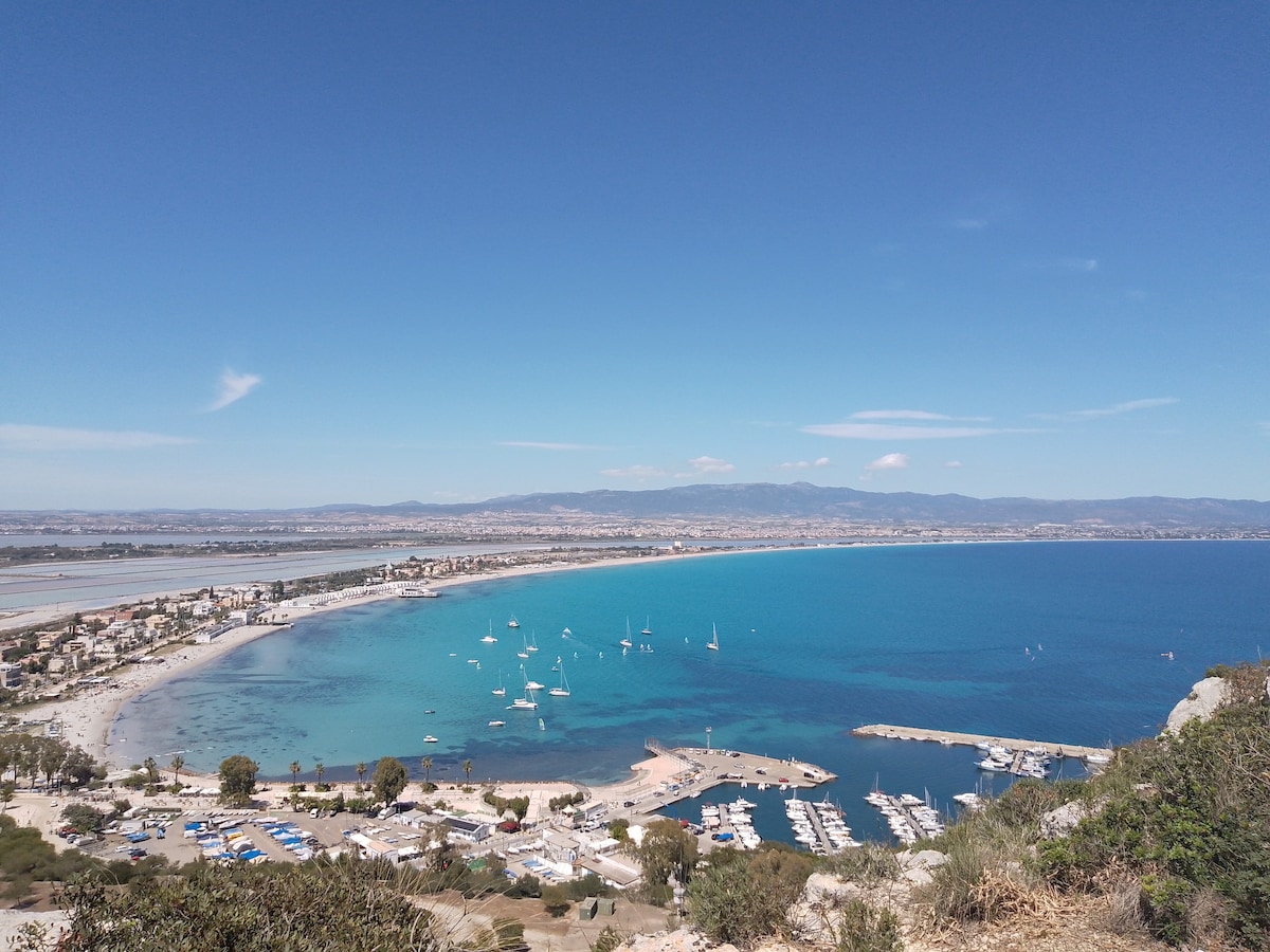 A panoramic view of a serene coastline features soft sandy beaches and a vibrant blue sea. Sailboats are anchored in the harbor, while the shoreline is lined with quaint buildings and lush greenery. The distant mountains create a tranquil backdrop, under a clear blue sky.