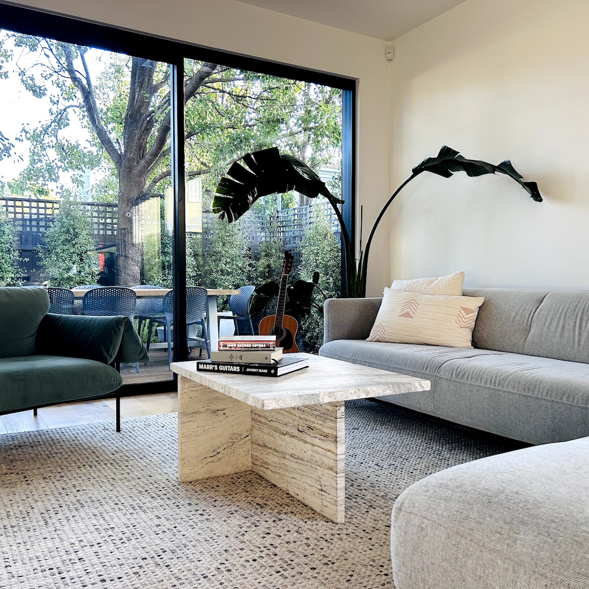 A modern living area features a light gray sofa and a deep green chair arranged around a unique marble coffee table. Large glass doors provide a view of the backyard, complemented by indoor plants that add a touch of nature to the space.